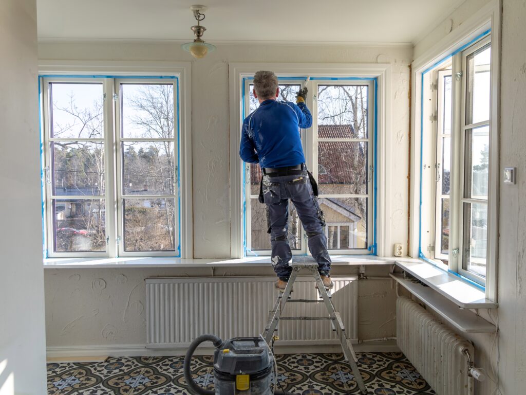 Ein Handwerker steht auf einer Leiter vor einem Fenster.
